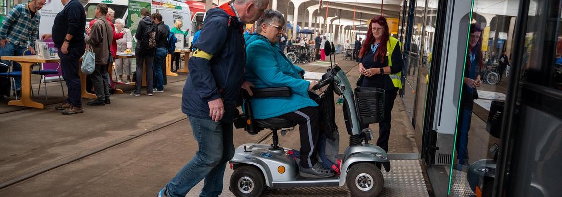 Person im elektrischen Rollstuhl und blinde Person steigen in Bahn ein mit Rampe