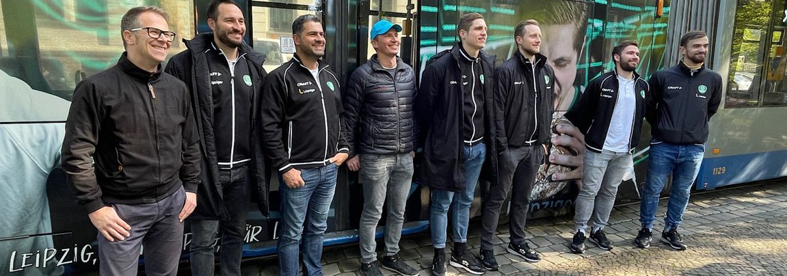 Vor einer Bahn auf den ein Handball-Motiv angebracht ist, steht das Team der SC DHfK-Handballer.