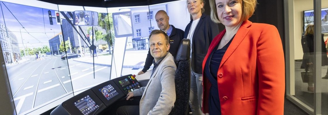 Man sieht das innere einer virtuellen Straßenbahnfhrerkabine. Statt Fenster sind dort Bildschirme zu sehen. Es liegt Schnee auf den Bildern, die über die Monitore zu sehen sind. Mehrere Personen sind in der Kabine.