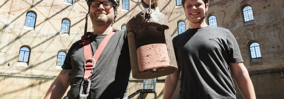 Zwei Männer stehen im alten Gasometer und halten lächelnd einen Nistkasten in die Kamera.