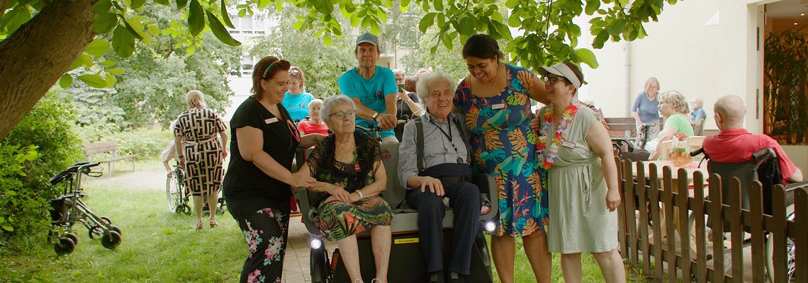 Mehrere Person stehen um ein Fahrrad herum. Auf dem Rad sitzen ältere Menschen.