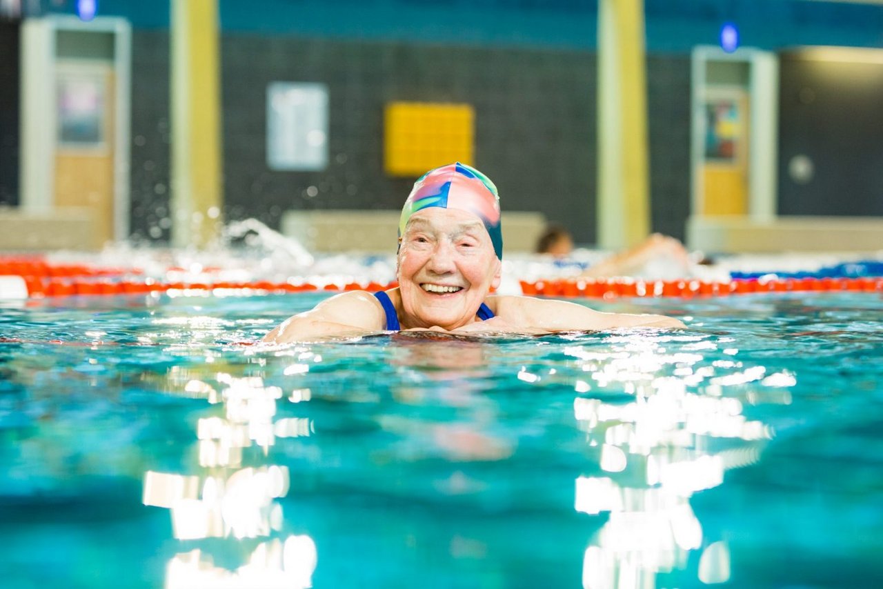 Eine ältere Frau schwimmt in einer Schwimmhalle lächelnd der Kamera entgegen. Sie trägt eine Badekappe.