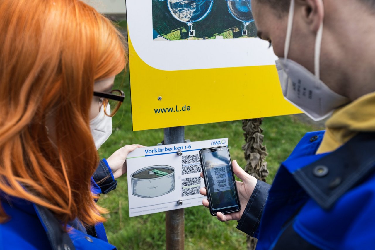 Zwei Personen stehen mit Handy vor einer Tafel auf der QR-Codes zu sehen sind.