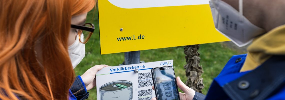 Zwei Personen stehen mit Handy vor einer Tafel auf der QR-Codes zu sehen sind.