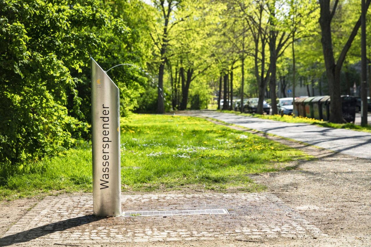 Ein Trinkbrunnen in der Totalen. Im Hintergrund sind Bäume und Wege in einem Park zu sehen.