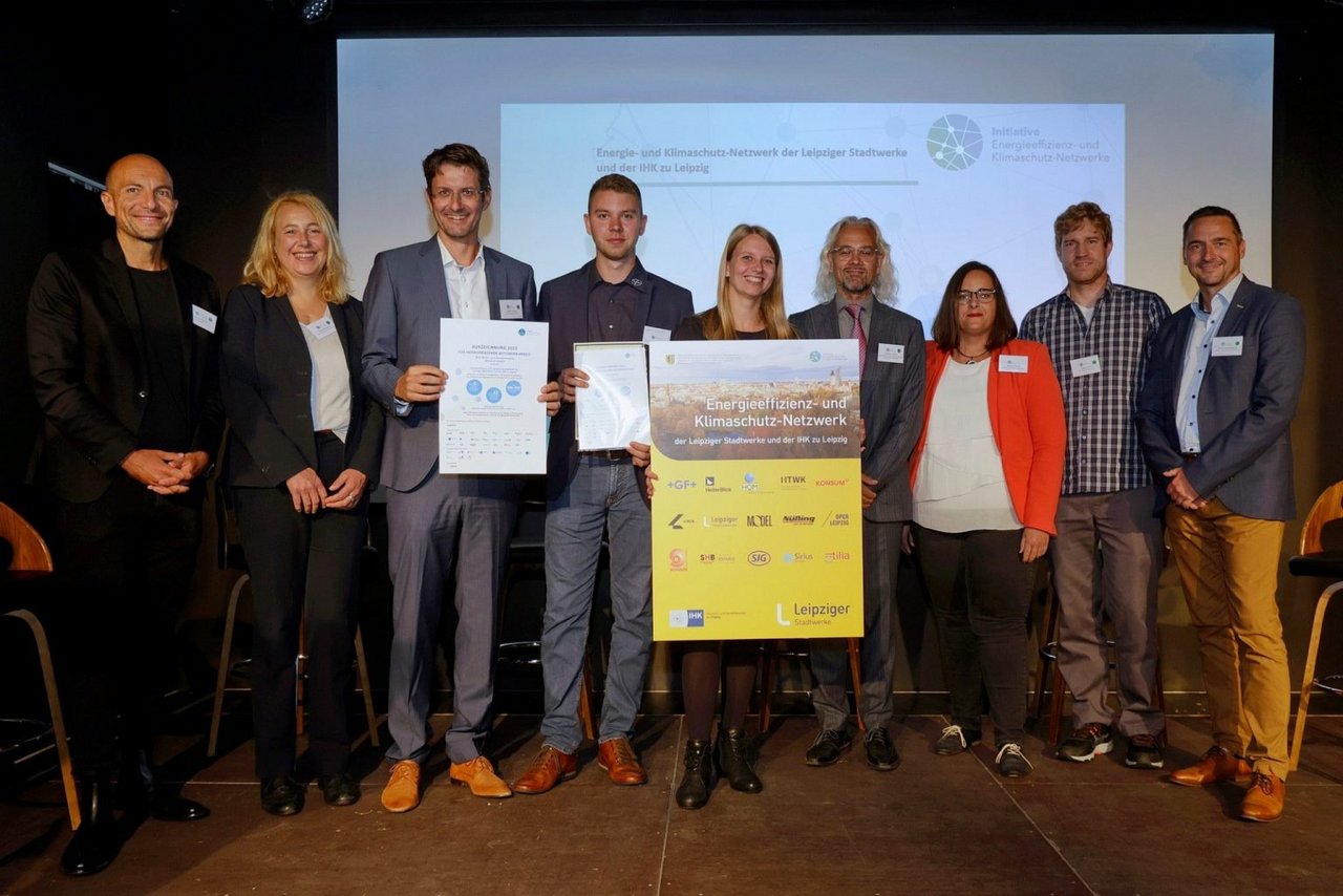 Gruppenfoto vom Netzwerktreffen mit Besichtigung des entstehenden Heizkraftwerks Süd am 1. September 2022 in Leipzig. Einige Personen halten eine Auszeichnung in die Höhe. Im Hintergrund ist eine Beamer-Leinwand. 