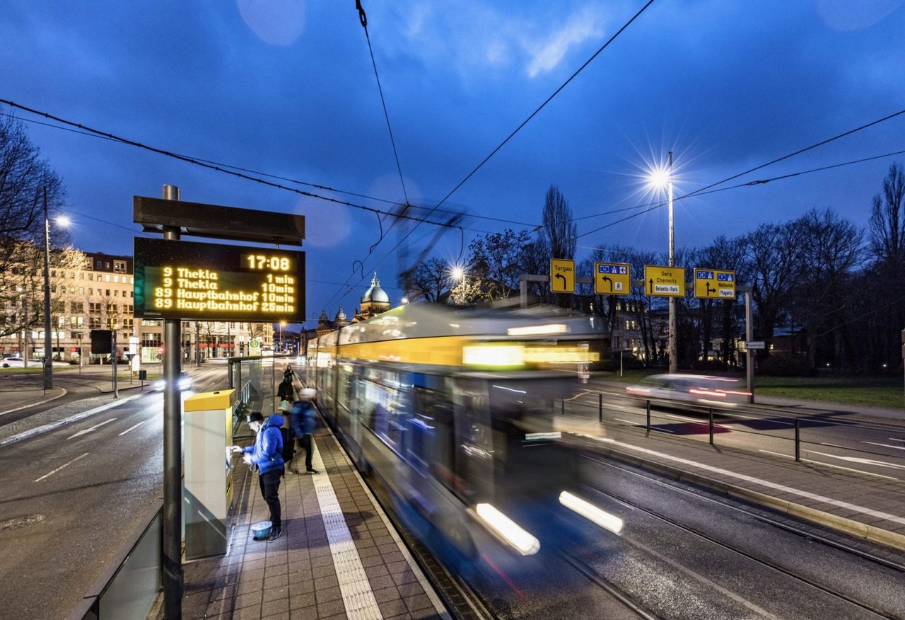 An einer Bahnhaltestelle fährt eine Bahn ab. Man sieht auch die digitale Echtzeitanzeige der Bahnabfahrten.