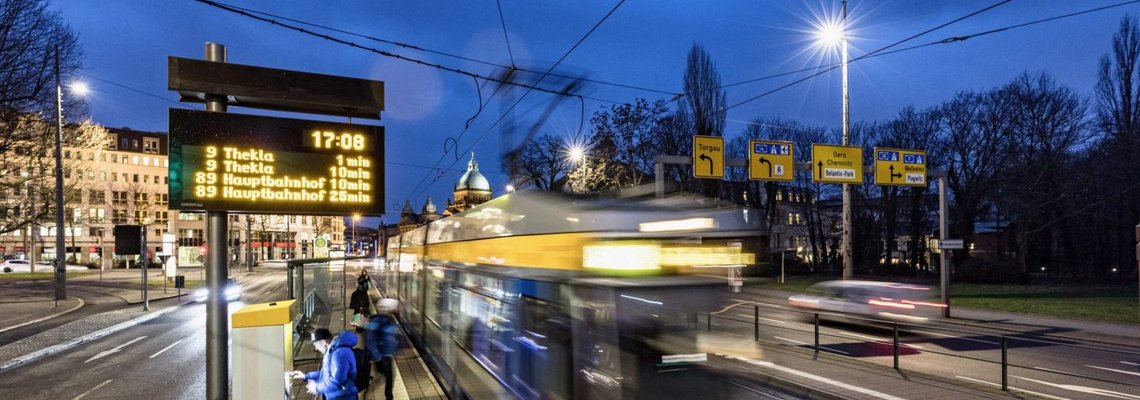 An einer Bahnhaltestelle fährt eine Bahn ab. Man sieht auch die digitale Echtzeitanzeige der Bahnabfahrten.