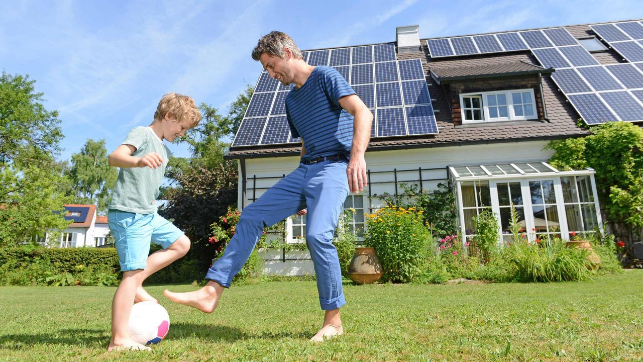 Vater und Sohn spielen im Garten Fußball, das Dach des Hauses im Hintergrund hat Photovoltaik-Panels. 