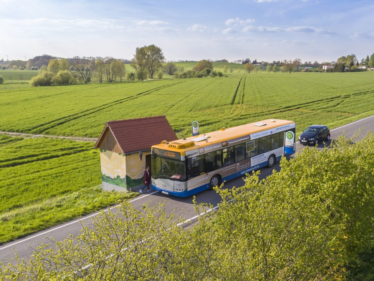 Ein Bus steht an einer Haltestelle. Um die Haltestelle herum sieht man Felder.