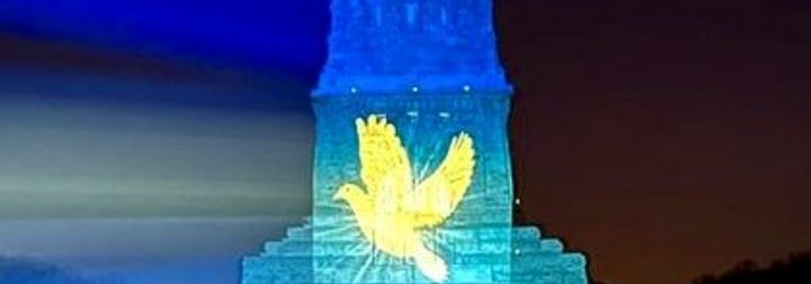 Das Völkerschlachtdenkmal wird in blau und gelb angestrahlt. Eine Friedenstaube wird darauf projeziert.