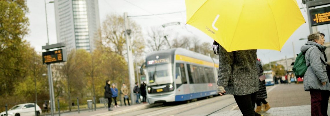 Eine Person steht mit gelben Gummistiefeln und einem gelbem Regenschirm an einer Haltestelle.