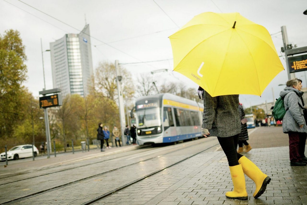Eine Person steht mit gelben Gummistiefeln und einem gelbem Regenschirm an einer Haltestelle.
