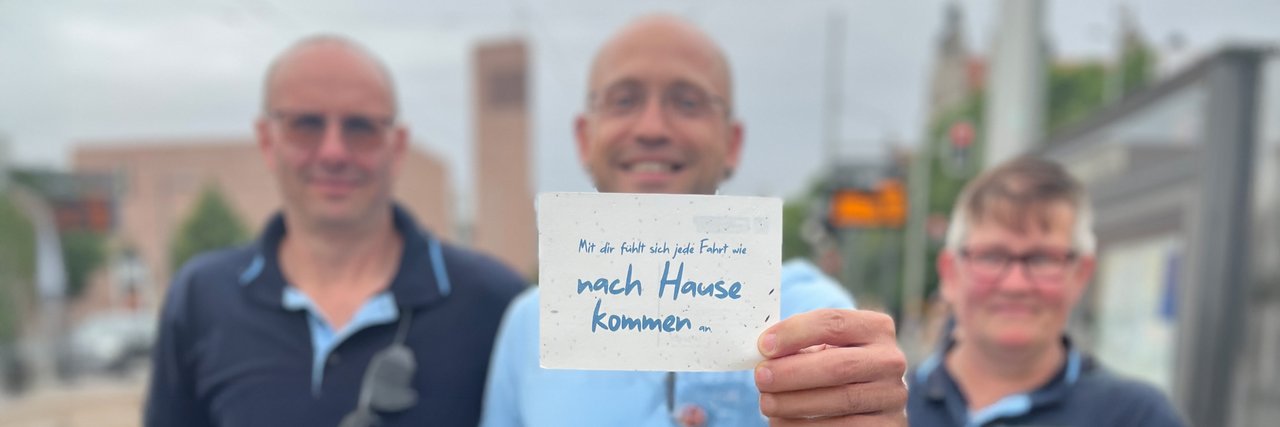 Drei Mitarbeitende der LVB halten eine Karte in die Kamera. darauf steht: Mit dir fühlt sich jede Fahrt an wie nach Hause kommen ...