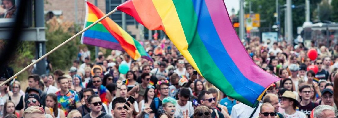 Mittig im Bild weht eine große Regenbogenflagge. Darunter stehen viele Menschen.